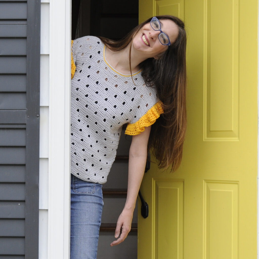 Mary Beth Cryan, a woman smiling leaning out of a door.  The door behind her is yellow.  Mary Beth is the indie crochet designer behind Crochet with Mary Beth a blog that features crochet garment and jewelry patterns and tips.
