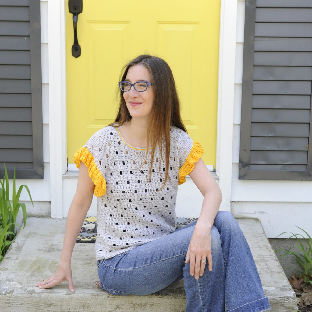 Mary Beth from Crochet with Mary Beth sitting on the steps in front of a yellow door modeling the Be Spotted Tee, a crochet garment made with Truboo yarn.