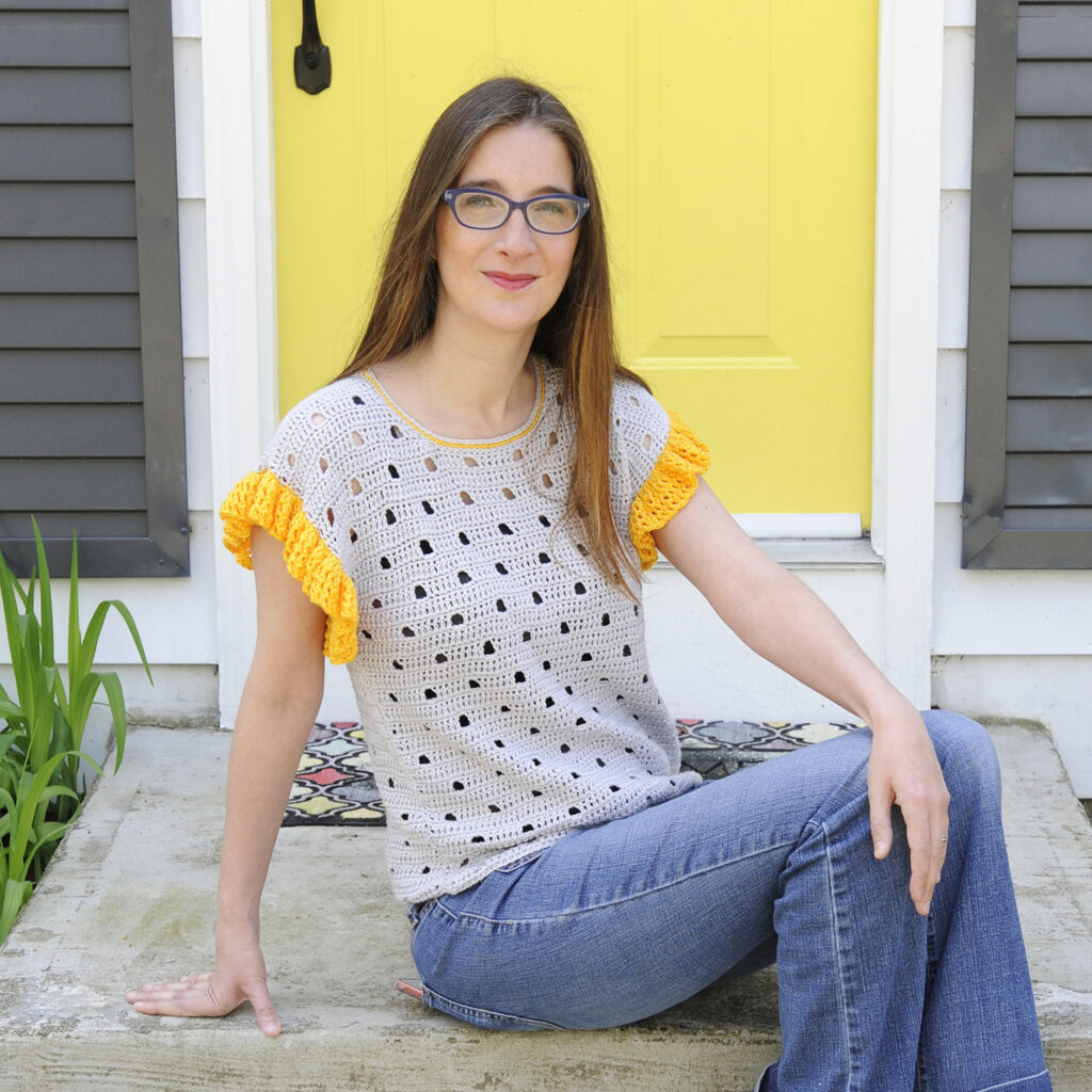 A woman sitting on the steps in front of a yellow door wearing a crochet top made out of Truboo Yarn, a sustainable bamboo yarn.