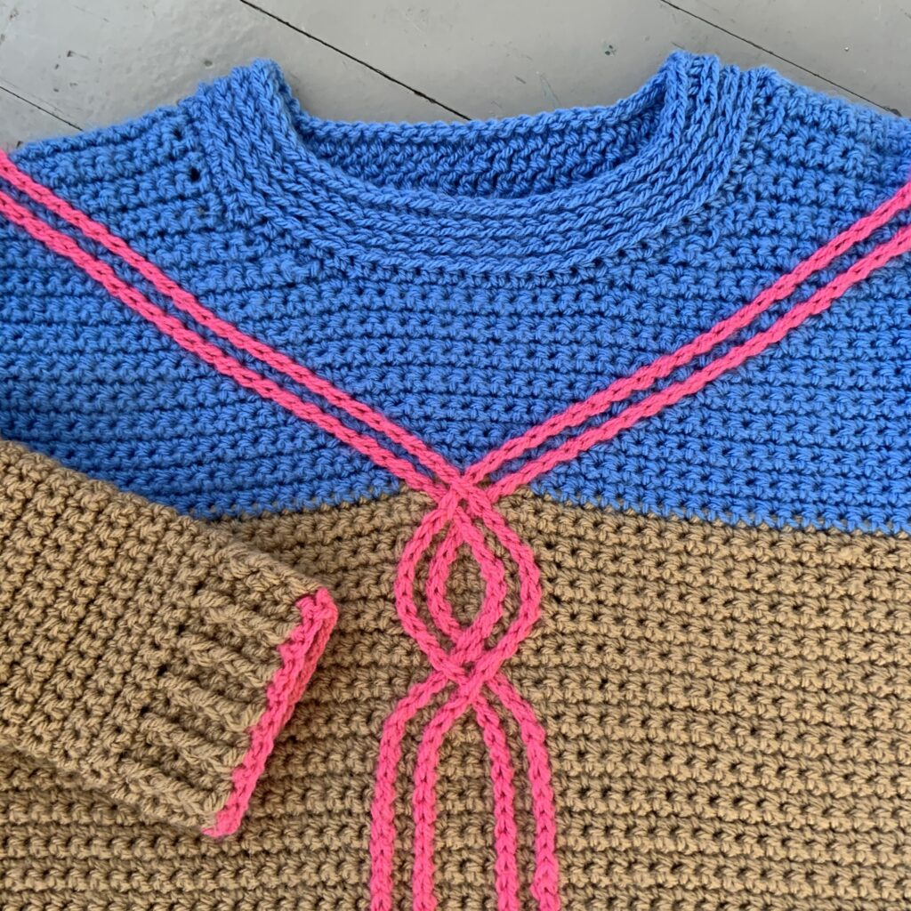Up close detail of a cable crochet sweater for women.  The sweater is blue and brown with a pink slip stitch surface crochet cable.