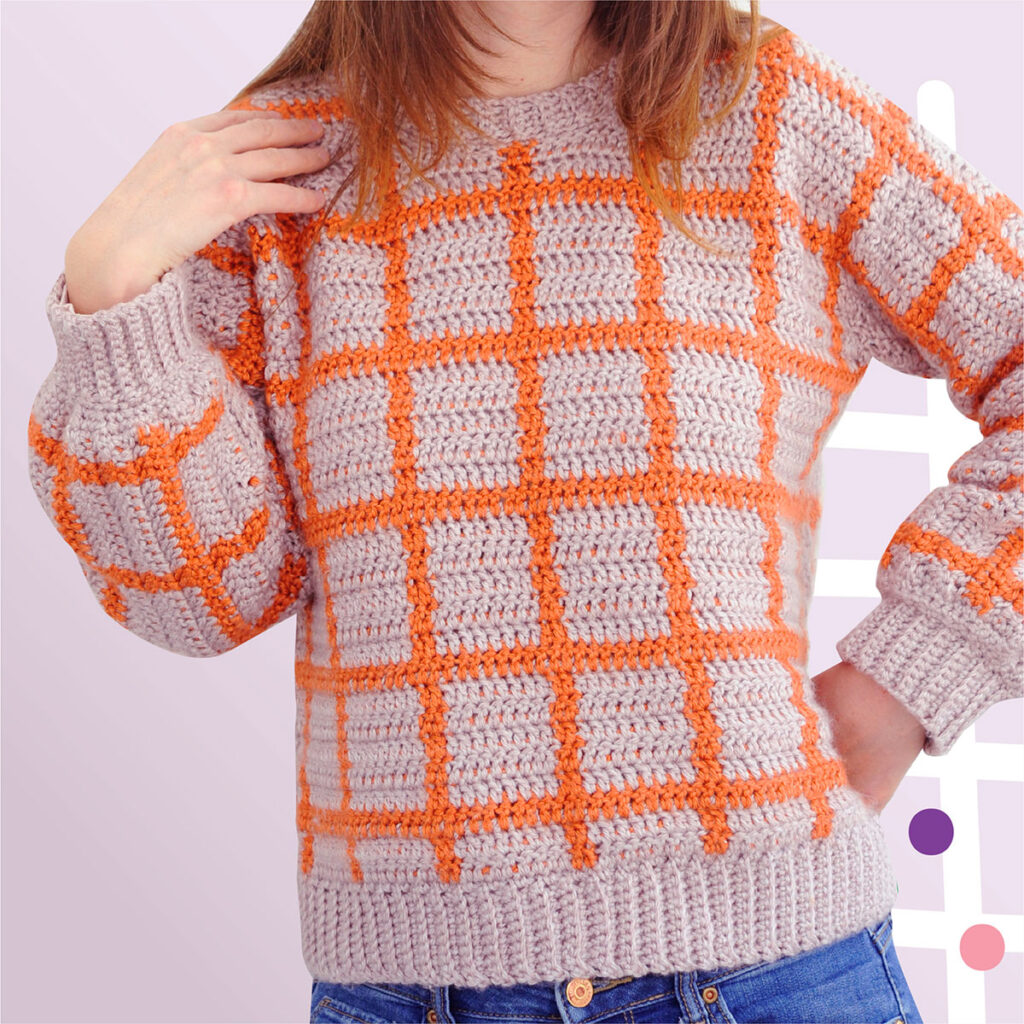 A crochet sweater pattern on a woman. The sweater is made out of worsted weight yarn. The colorful pattern is made out of a grid of orange and light purple tapestry crochet.