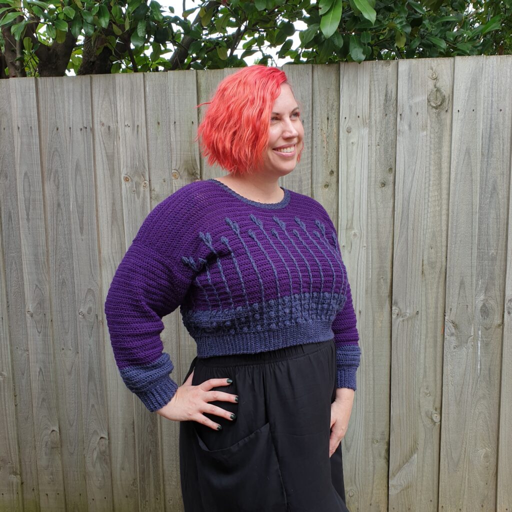 A woman wearing the cropped version of the Best Buds Sweater, a modern crochet sweater pattern. The top is purple and dark blue with a floral cable pattern.