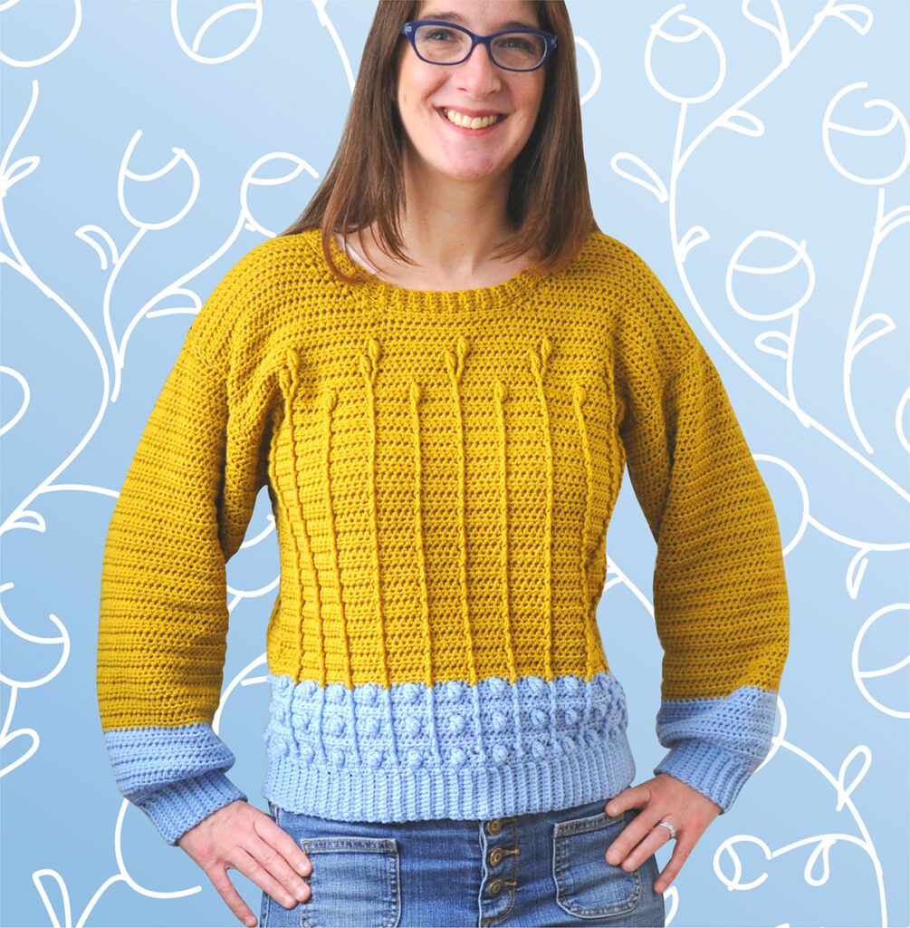 A woman wearing a crochet sweater. The sweater is mustard and blue and features bobbles and floral cables.