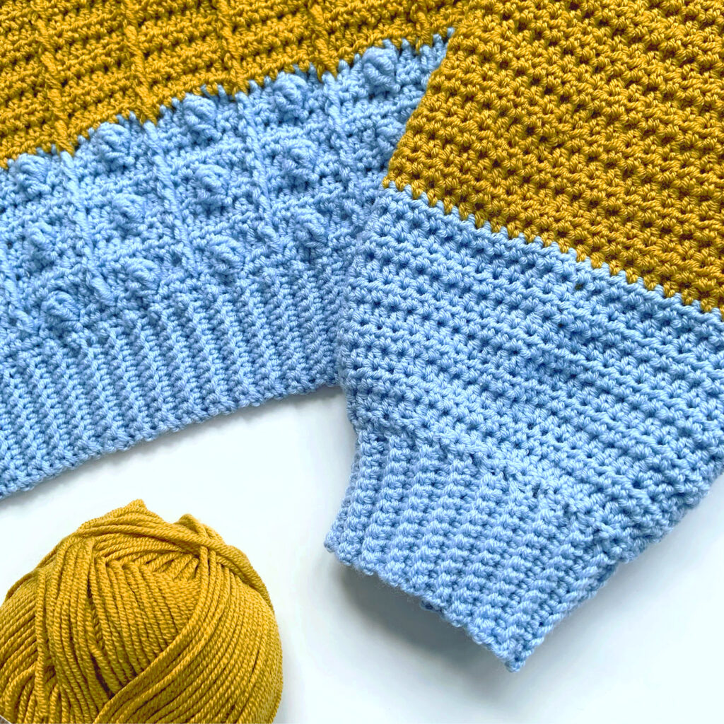 Clos-up of a modern crochet sweater featuring a blue bobble and cable pattern that turns into a gold floral stitch pattern.