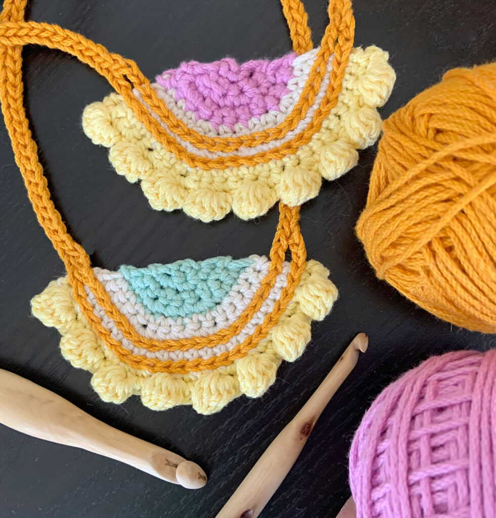 Two pieces of crochet jewelry made from the crochet necklace pattern Eight Bobble Necklace. The necklaces are lilac, white, gold, and yellow. Two wood hooks and two balls of yarn are also shown.