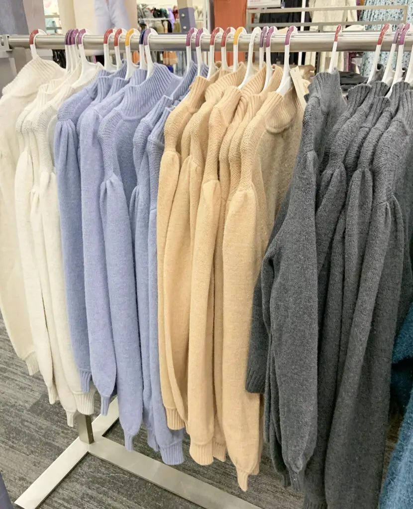 A rack of sweaters at a store in the colors white, light periwinkle, beige, and gray.
