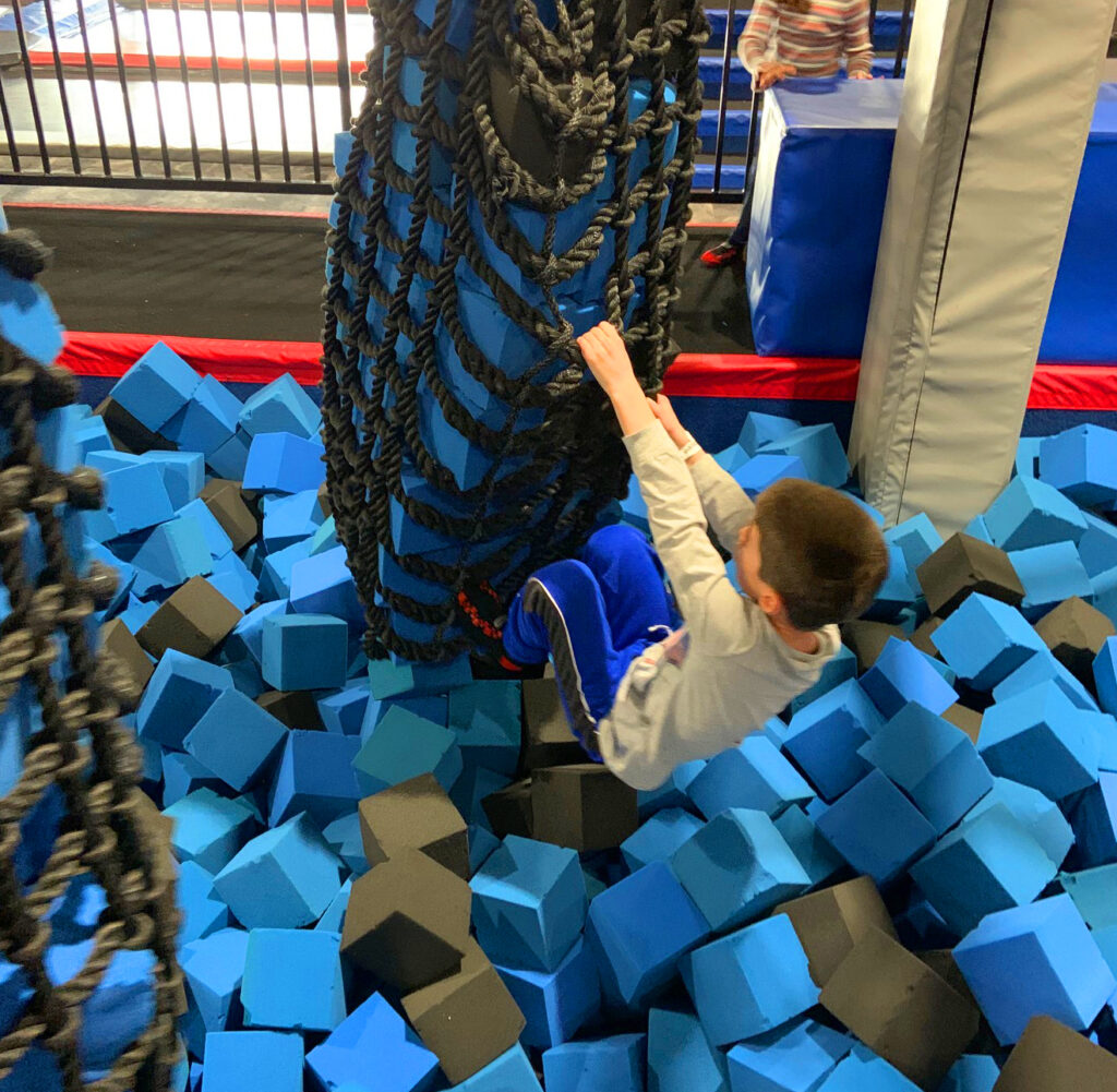 A boy swinging on a rope ladder at a trampoline park.