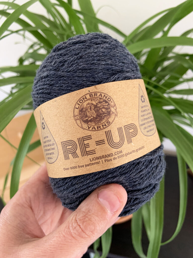 An image of a woman's hand holding a skein of Lion Brand's sustainable yarn Re-Up in dark blue.