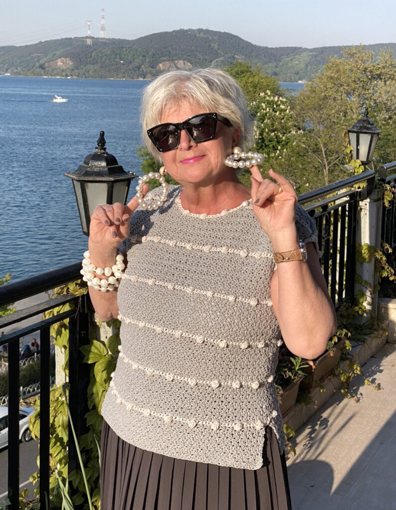 A woman wearing the Lean Into Bobbles crochet summer top pattern.  The bobbles are white against a gray crochet background.