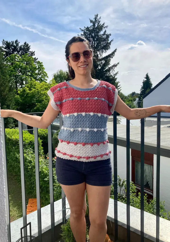 A woman wearing the Lean Into Bobbles crochet t shirt in pink, gray and white standing on porch on a sunny day.