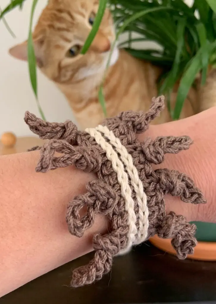 A cat peaking from behind a plant at a crochet diy bracelet.  The crochet bracelet is brown and features a knotted border and slip stitch crochet.