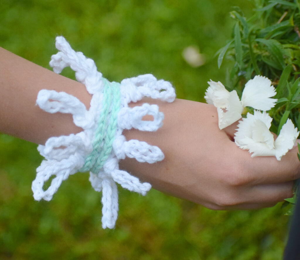 A woman's hand holding flowers wearing a white and mint crochet bracelet featuring a knotted crochet border and slip stitch crochet.
