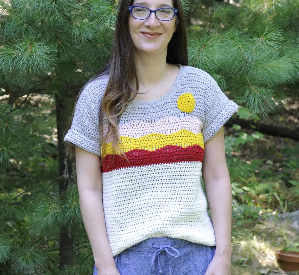A woman wearing a crochet t shirt depicting a desert scene.  The colors of the shirt are red, yellow, pink, white and gray.