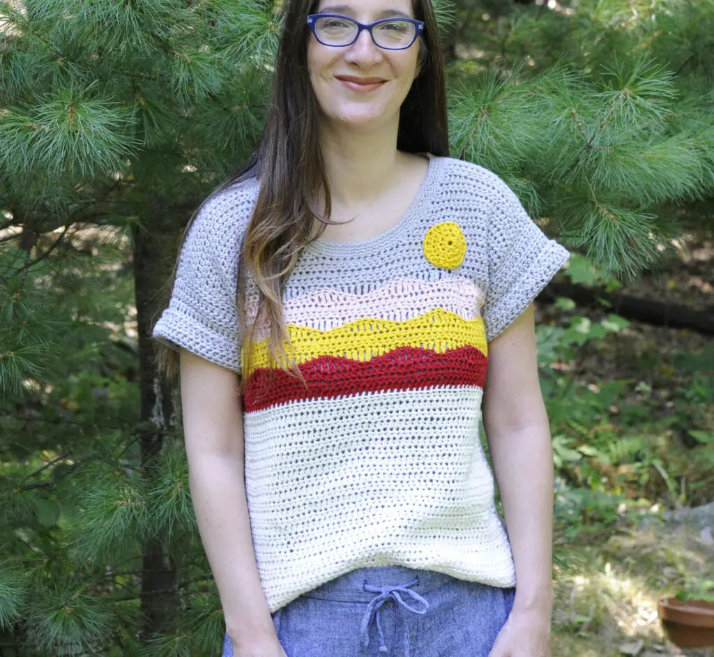 A woman wearing a crochet t shirt depicting a desert scene. The colors of the shirt are red, yellow, pink, white and gray.