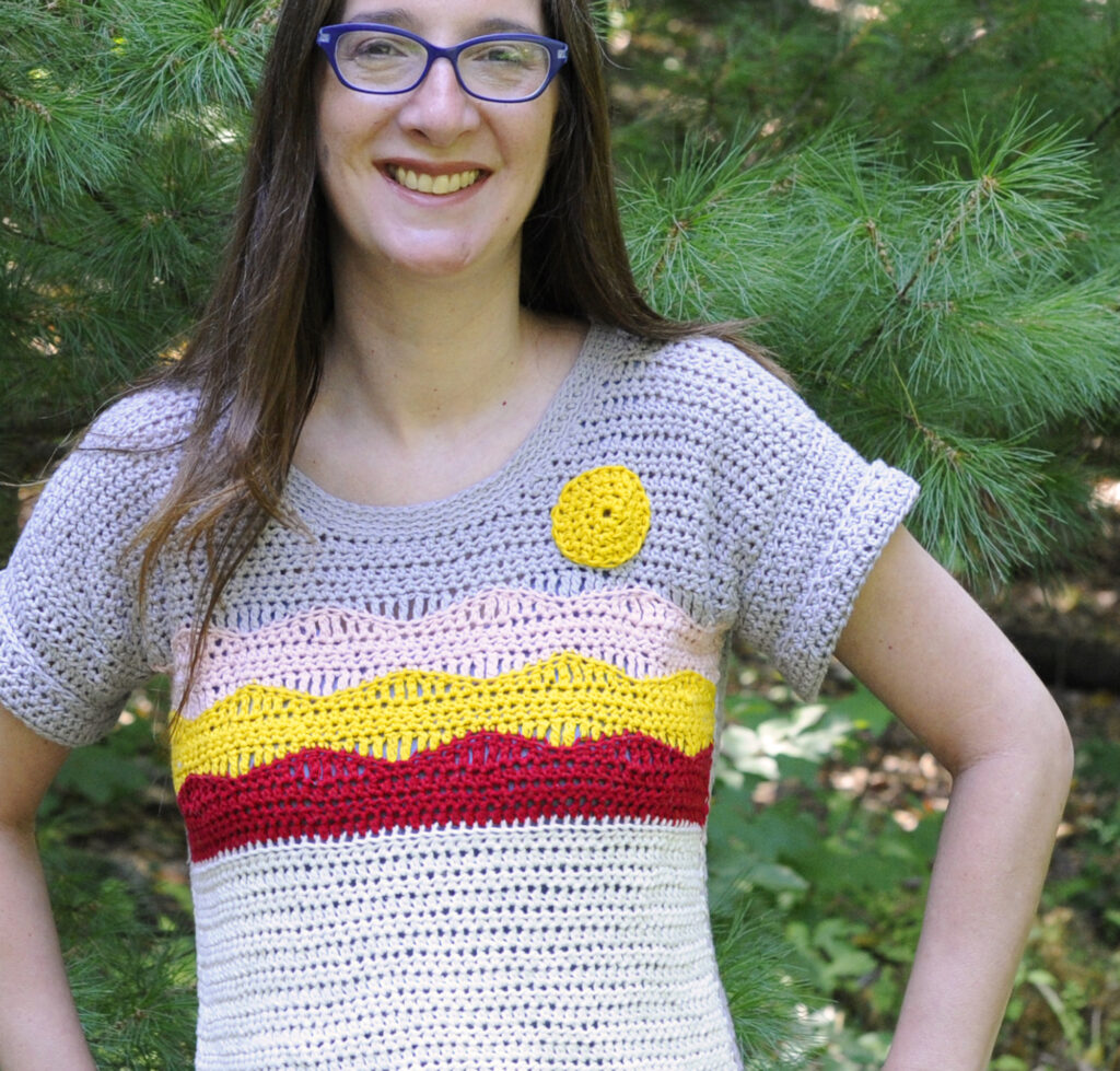 A woman wearing a crochet top pattern.  The top has a picture of a mountain on it created in crochet stitches.